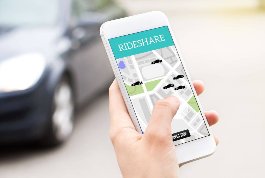 Hand holding mobile phone with rideshare app on screen with dark-colored car in background.